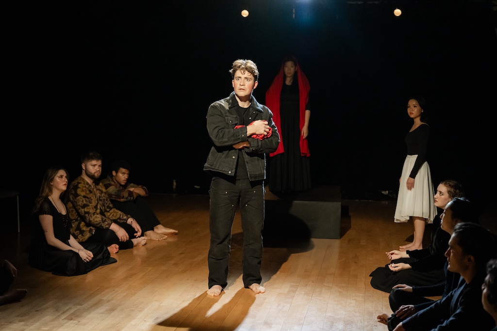 Actor stands on stage holding a crumpled bright red cape in his arms while other actors sit cross-legged on either side of him. Another actor wearing a bright red cape stands on a black riser directly behind him.